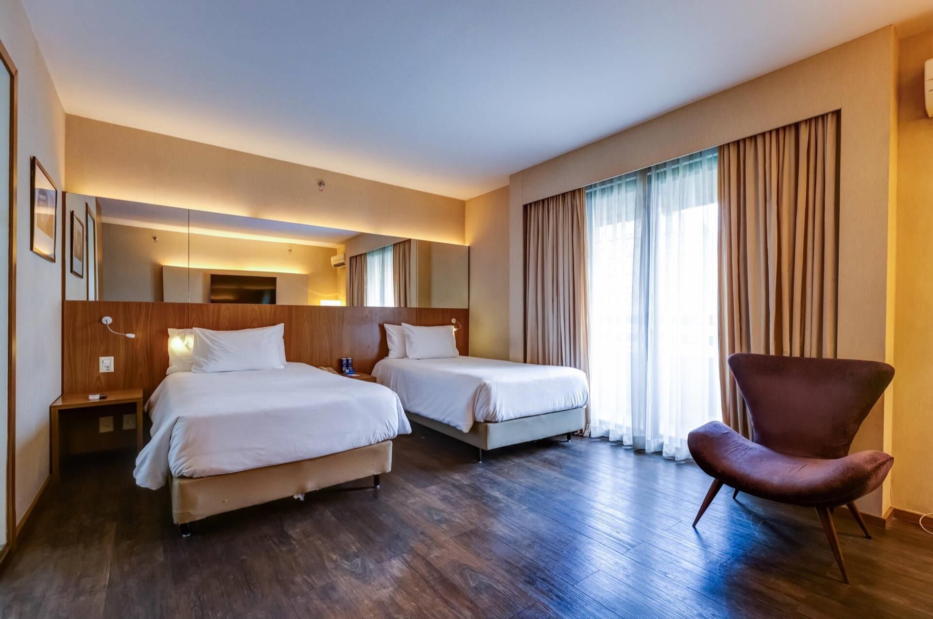 Une chambre d’hôtel avec deux lits, une chaise, un miroir et une grande fenêtre avec des rideaux.