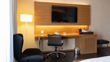 Een hotelkamer met een houten bureau, een zwarte bureaustoel, een aan de muur bevestigde televisie, een nachtkastje en een bed.