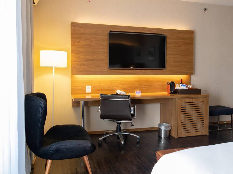 Een hotelkamer met een houten bureau, een zwarte bureaustoel, een aan de muur bevestigde televisie, een nachtkastje en een bed.