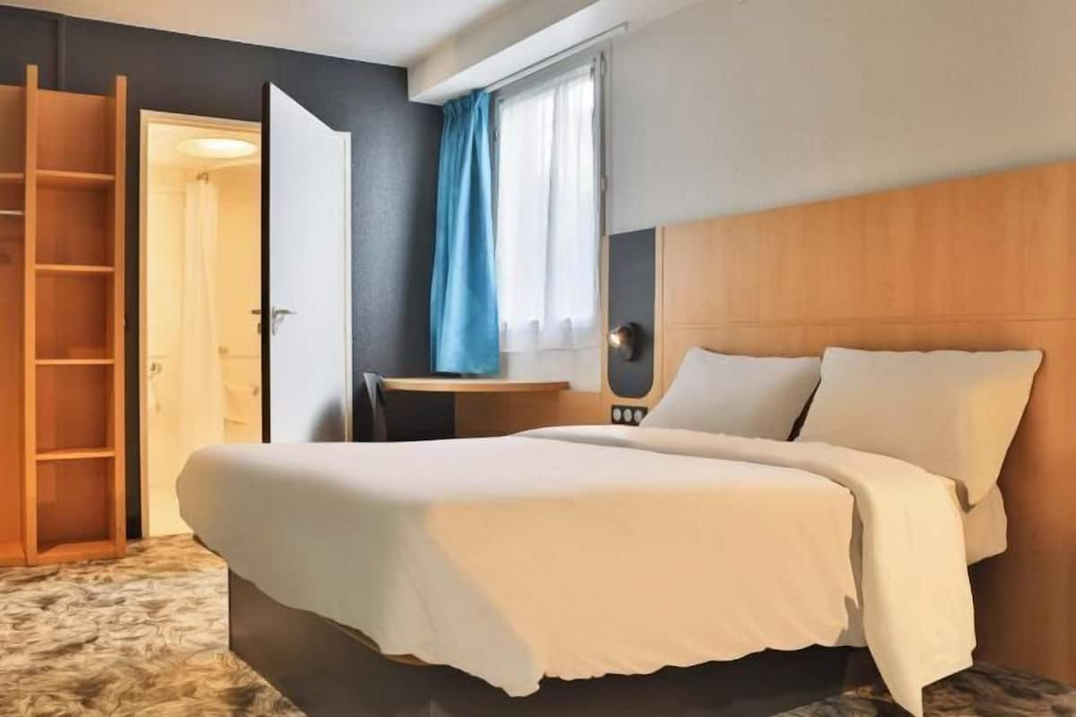 Une chambre d’hôtel avec un lit, un bureau, une chaise, une échelle en bois et une fenêtre avec des rideaux.