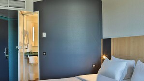 Habitación de hotel con cama, mesita de noche, luz empotrada en la pared y puerta abierta a un baño.