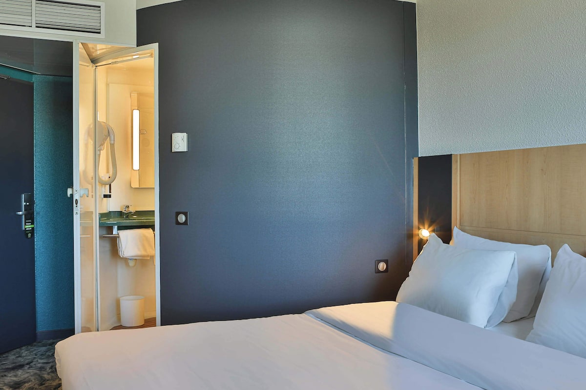 Une chambre d’hôtel avec un lit, une table de chevet, une lampe fixée au mur et une porte ouverte donnant sur une salle de bain.