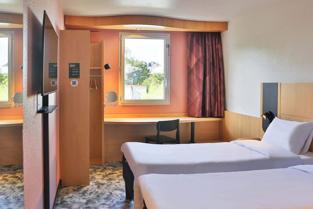 Une chambre d’hôtel avec deux lits, un bureau, une chaise, une télévision et une fenêtre avec des rideaux.