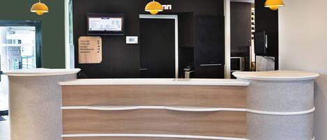 Un mostrador de recepción de diseño moderno, con acabado de madera y forma curvada. Dos lámparas colgantes están suspendidas sobre el mostrador, y una pantalla digital muestra información en la pared de fondo.