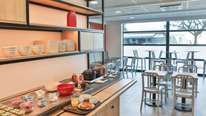 Una barra de desayuno con diversas opciones de comida y utensilios, rodeada de sillas y mesas en una habitación luminosa con ventanas grandes.