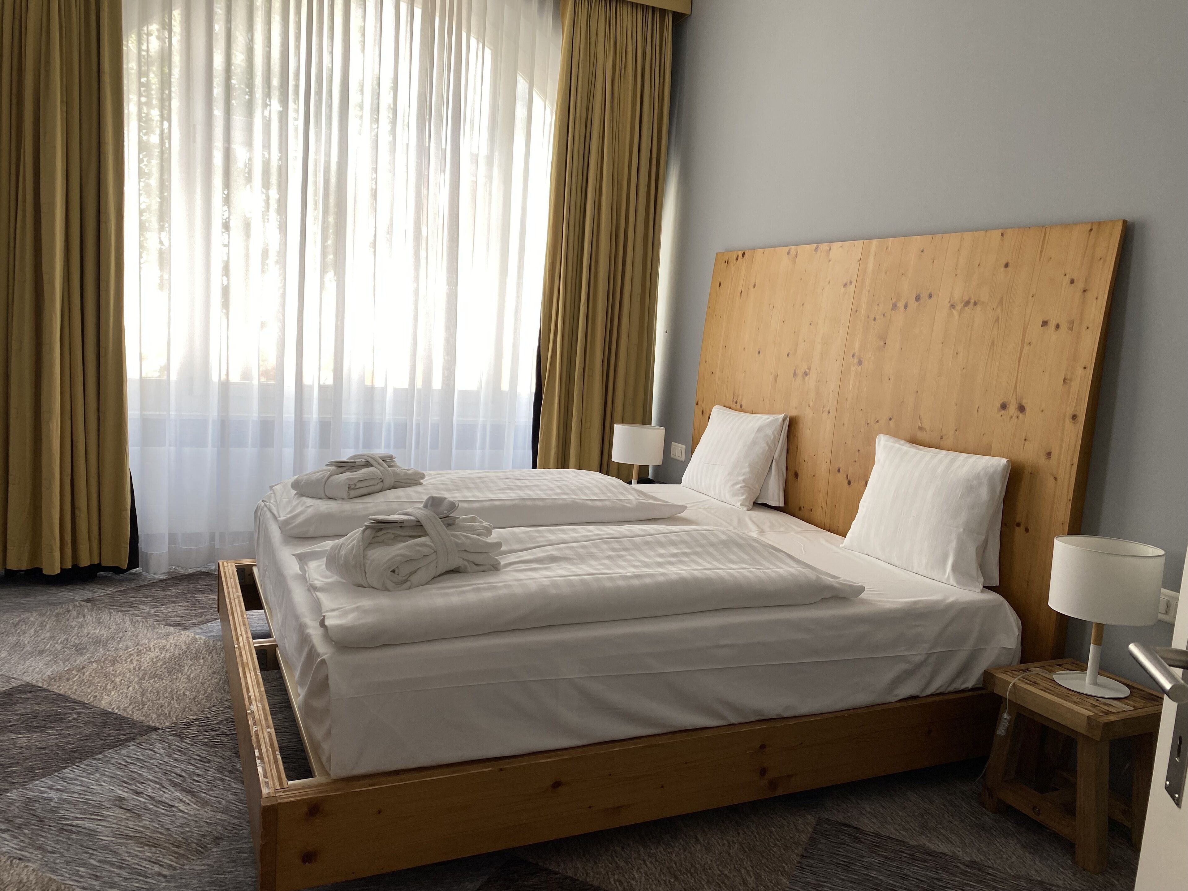 A hotel room with a wooden bed frame, white linens, and a view through sheer curtains.