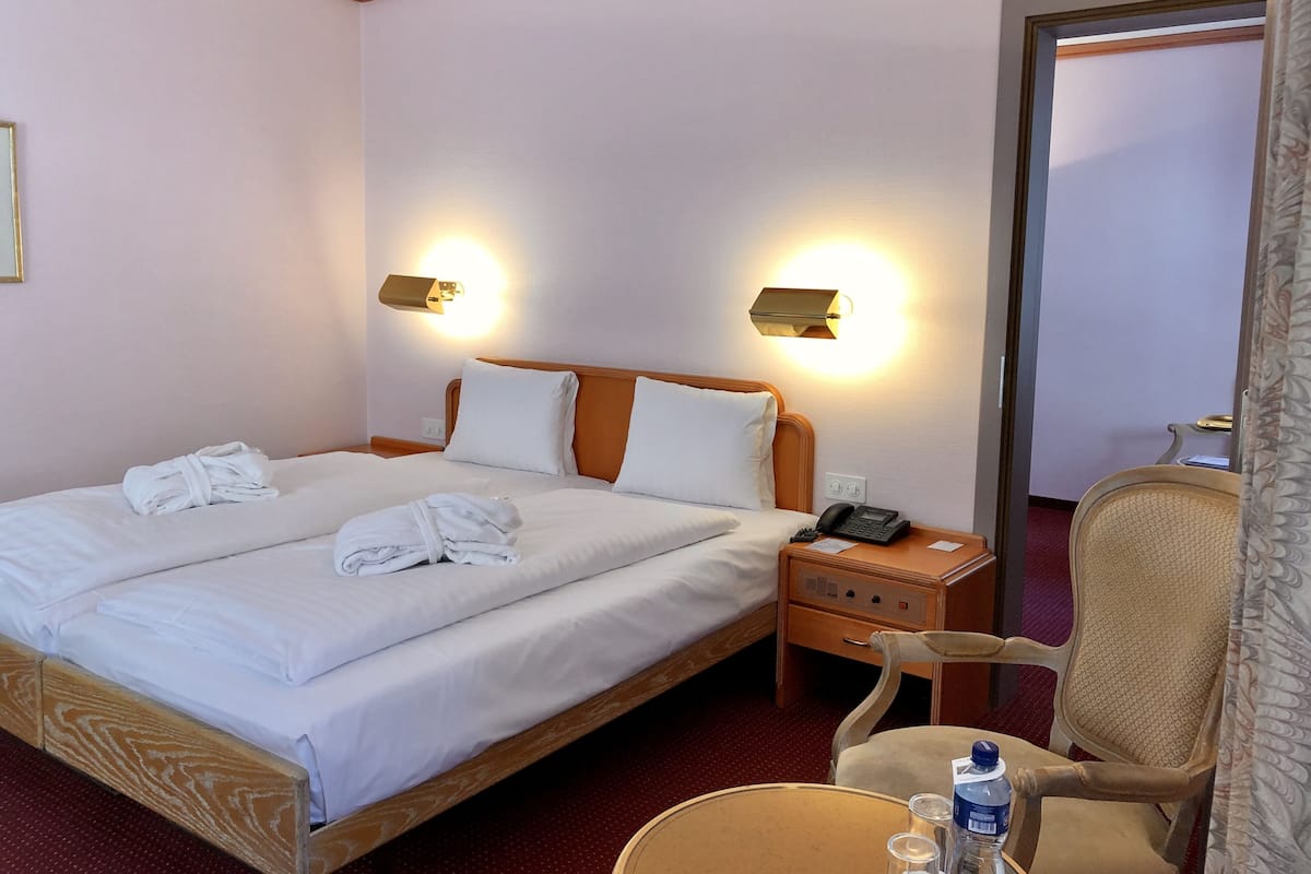 Hotelkamer met een tweepersoonsbed, twee bedlampen, een bedtelefoon, een stoel en een klein tafeltje met een fles en glaasjes.