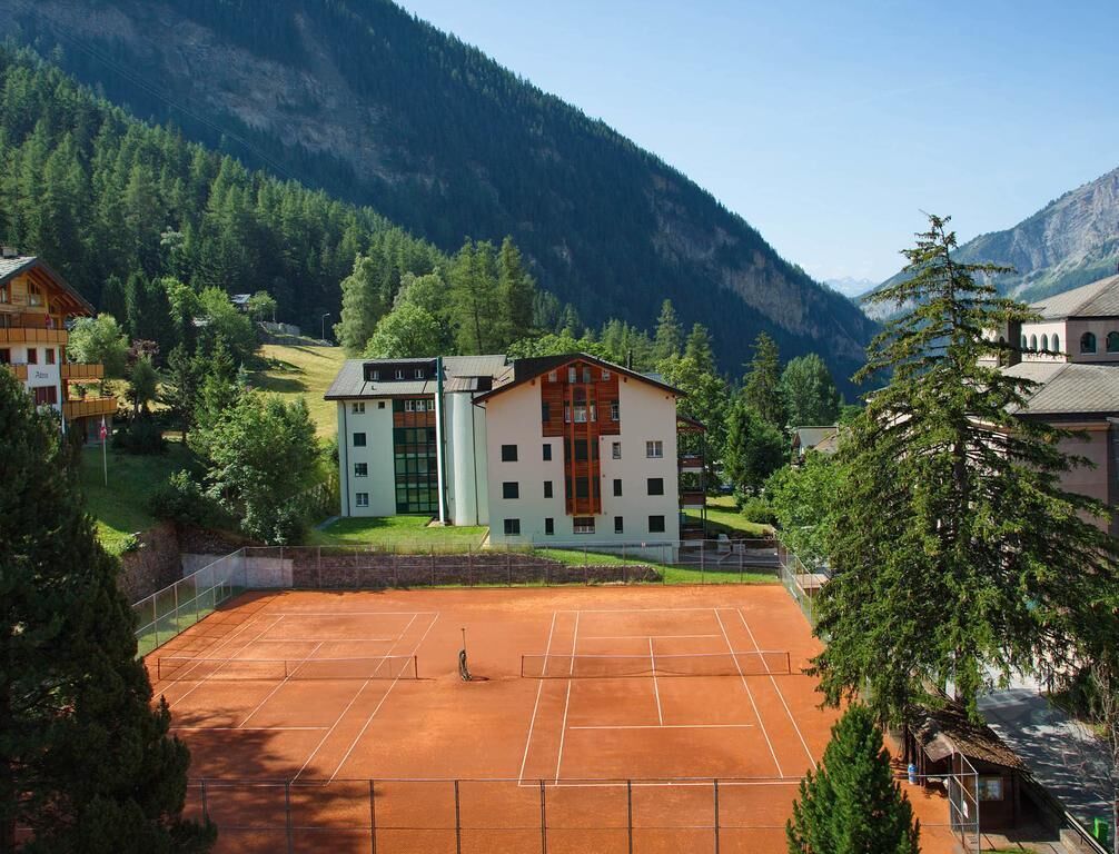 A tennis court surrounded by buildings with a mountainous backdrop.