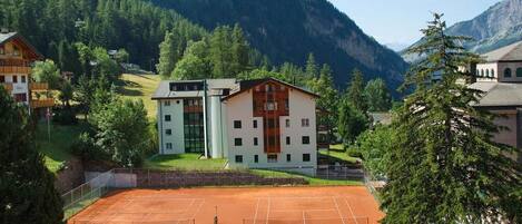 A tennis court surrounded by buildings with a mountainous backdrop.