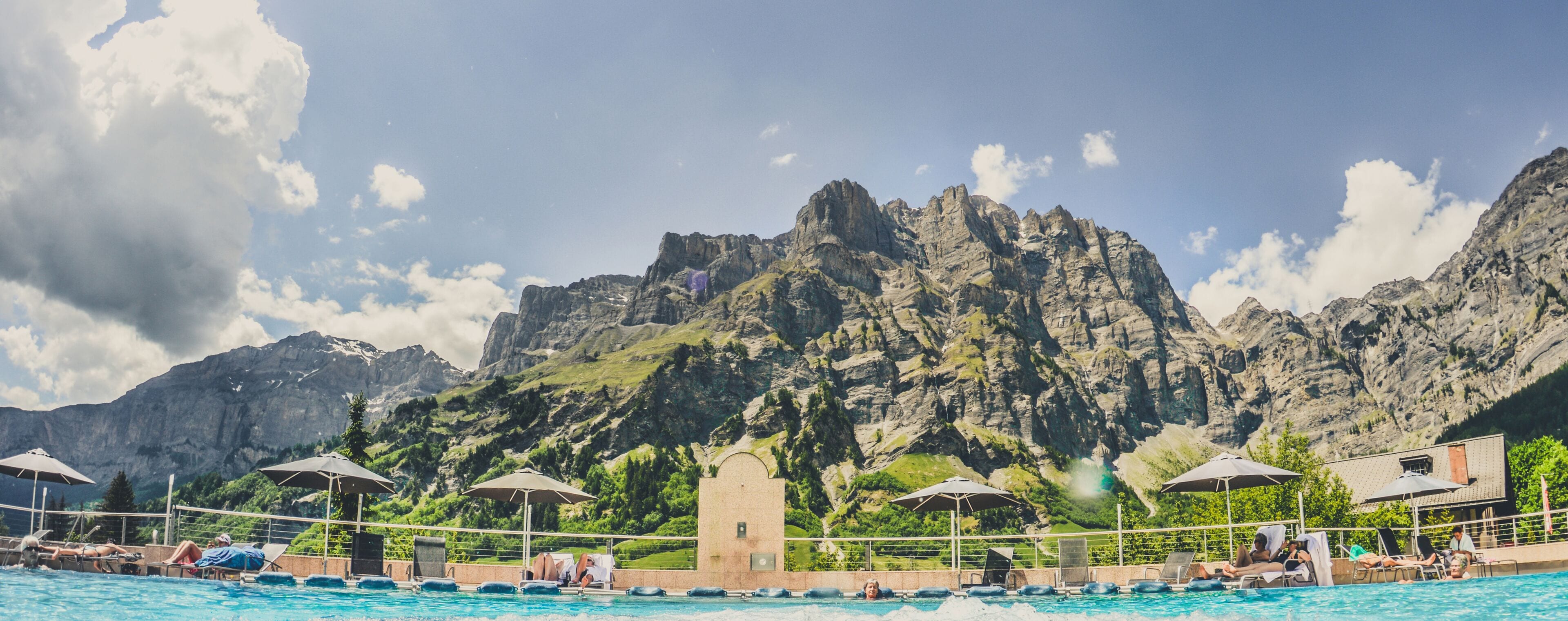 Una piscina con lettini e ombrelloni, circondata da montagne e un cielo sereno.