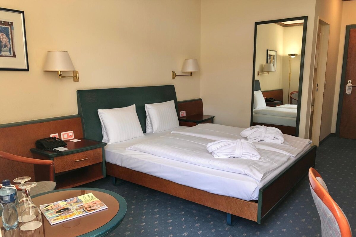 Een hotelkamer met een bed, nachtkastjes, een ronde tafel met een tijdschrift, een stoel, een spiegel en een wandlamp.