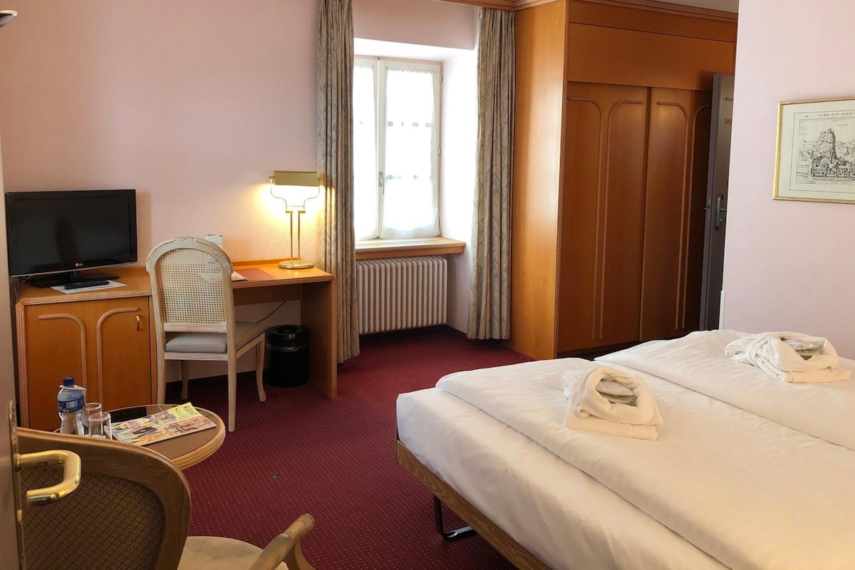 Een hotelkamer met een bed, een bureau met lamp, een televisie, een stoel, een kledingkast en een raam met gordijnen.