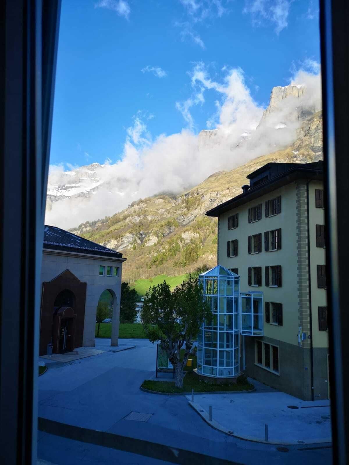 A view of a building with a glass structure in the foreground, a stone building with arches, and a mountain with clouds in the background.