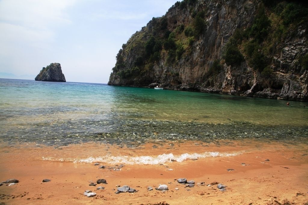 Una spiaggia con acque limpide, uno sperone roccioso e un'isola in lontananza.