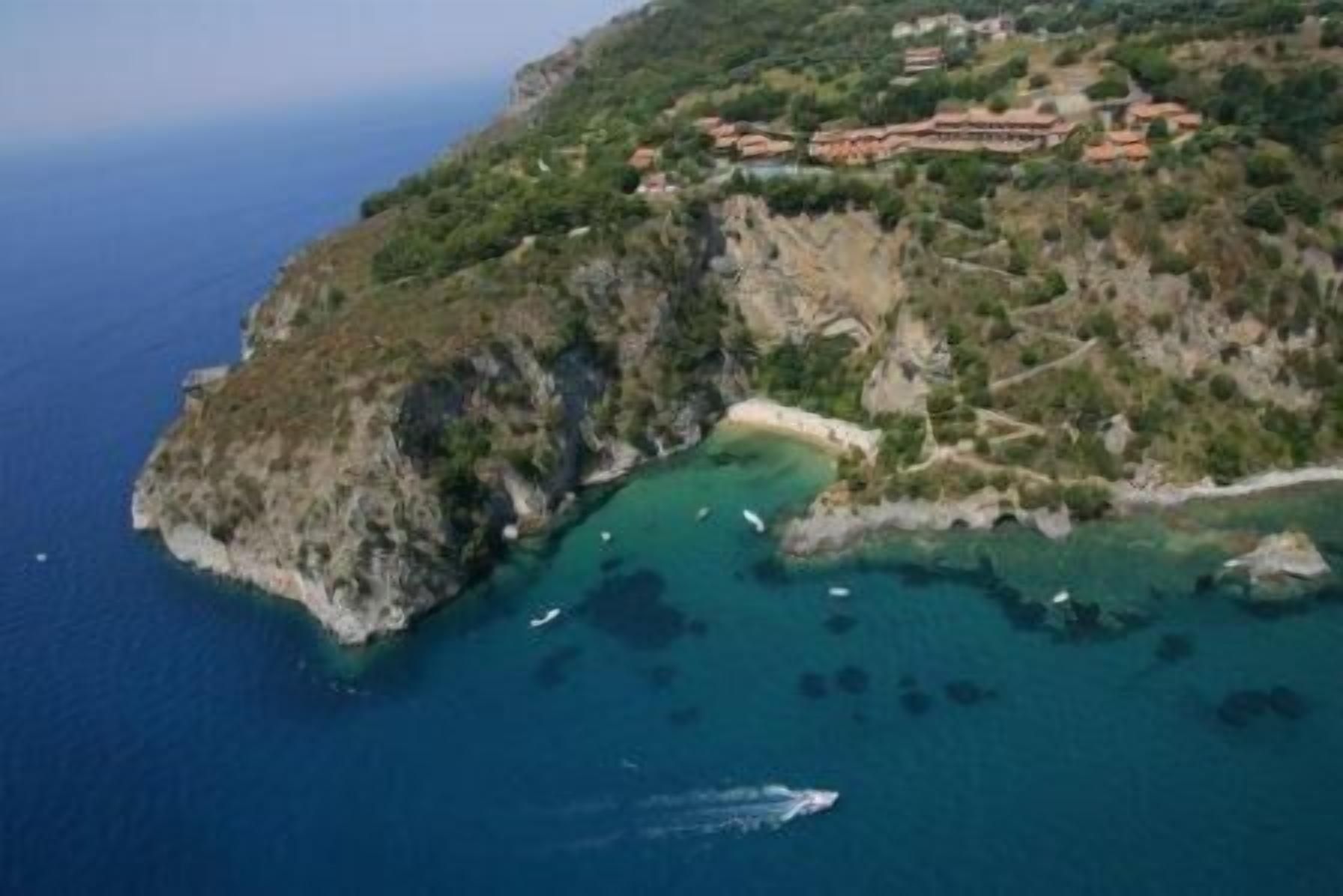 An aerial view of a coastal area with a cluster of buildings on a hillside, surrounded by clear blue waters and rocky terrain.