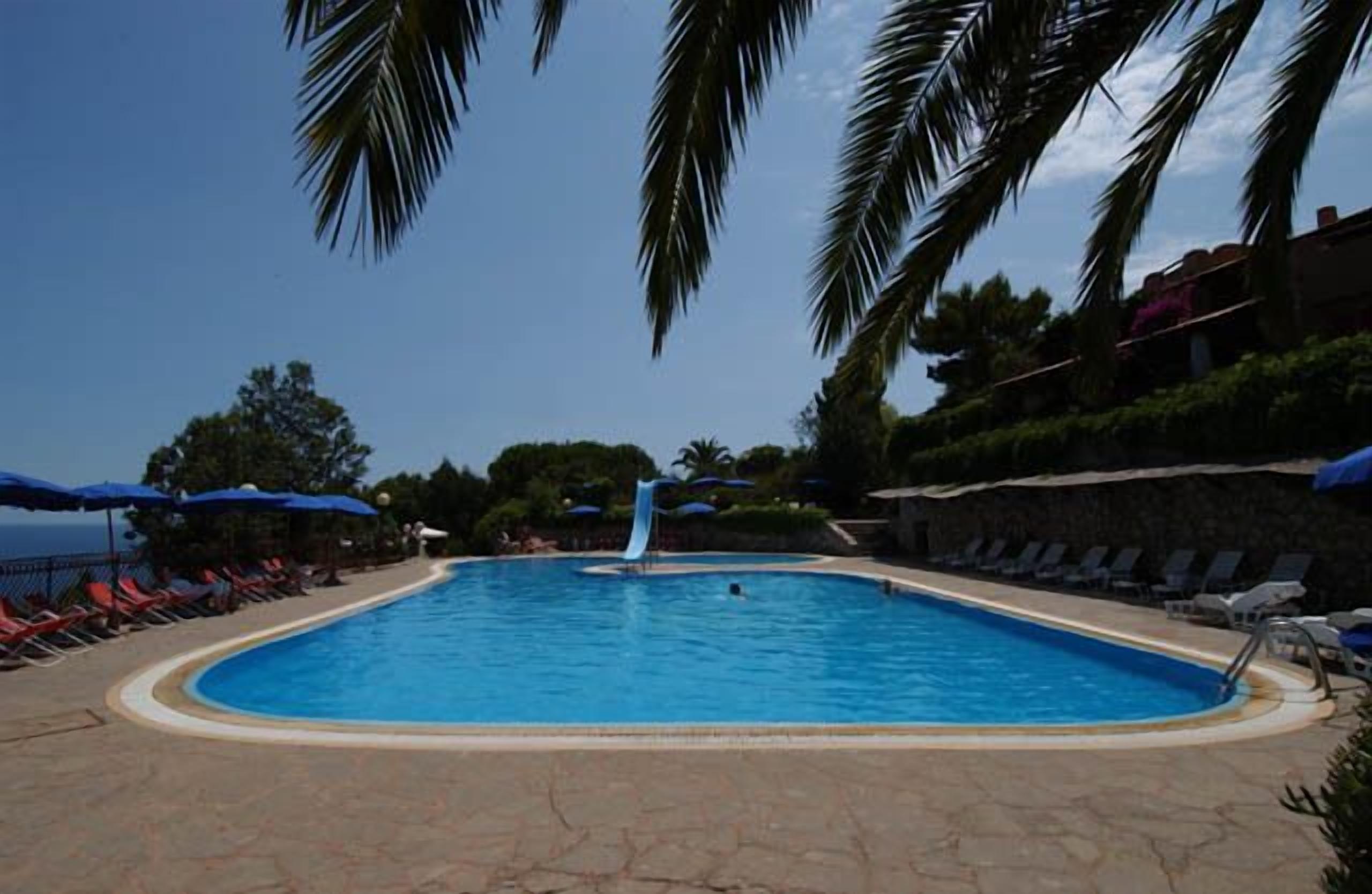A swimming pool with a slide, surrounded by lounge chairs and umbrellas, with palm trees and a clear sky in the background.