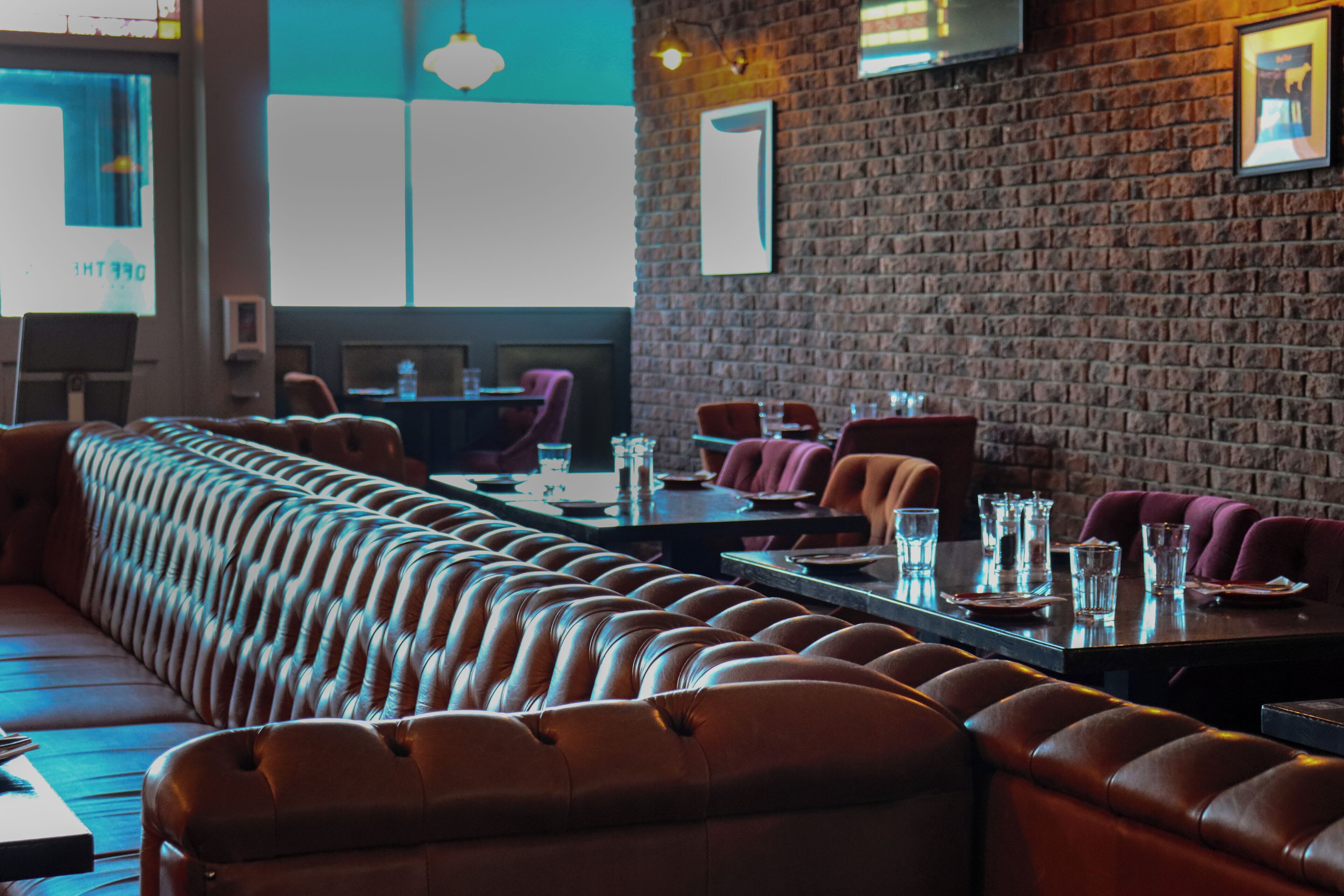 A restaurant with a long leather couch, brick walls, and tables set for dining.