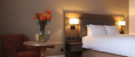 A hotel room with a bed, a nightstand, a table with a vase of flowers, and a chair.