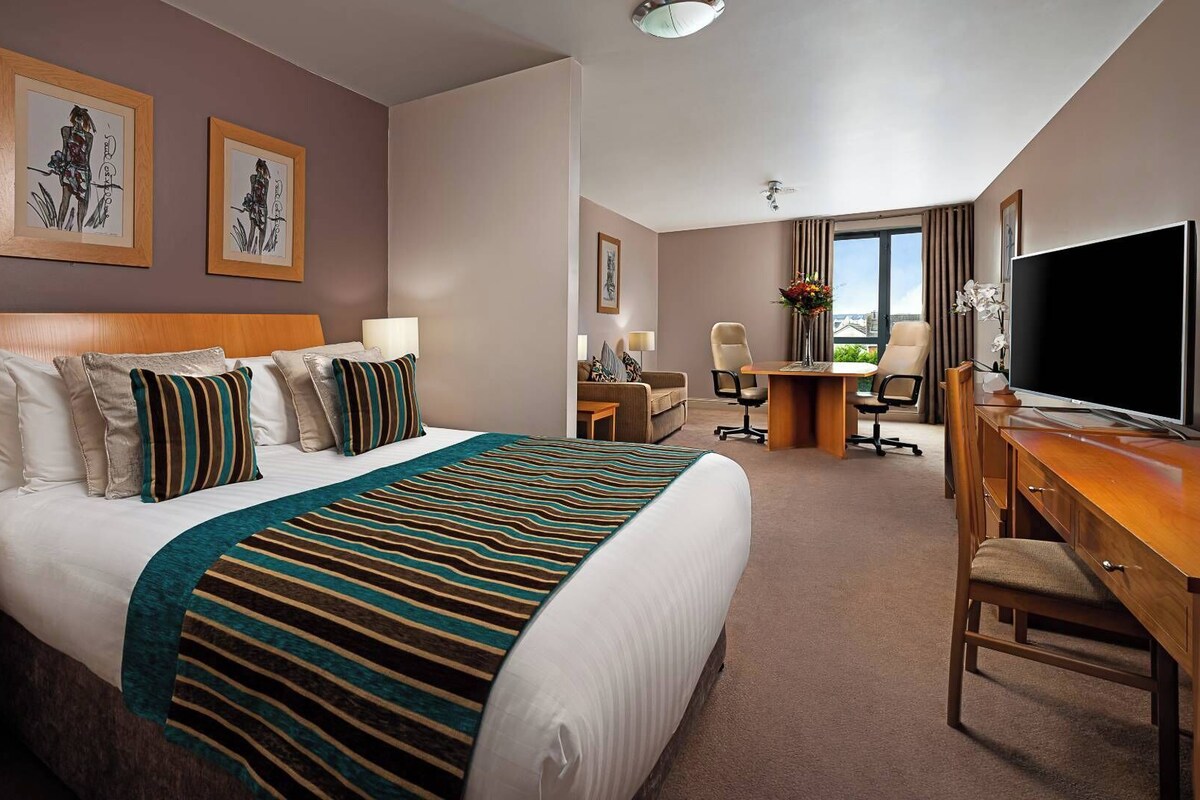 A hotel room with a large bed, a desk, a TV, and a view of the city.