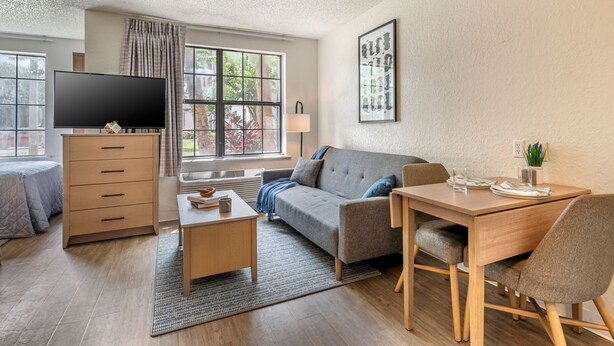 Una sala moderna con sofá, una mesa de centro, televisor sobre un mueble de madera, una mesa de comedor con sillas y una ventana con cortinas.