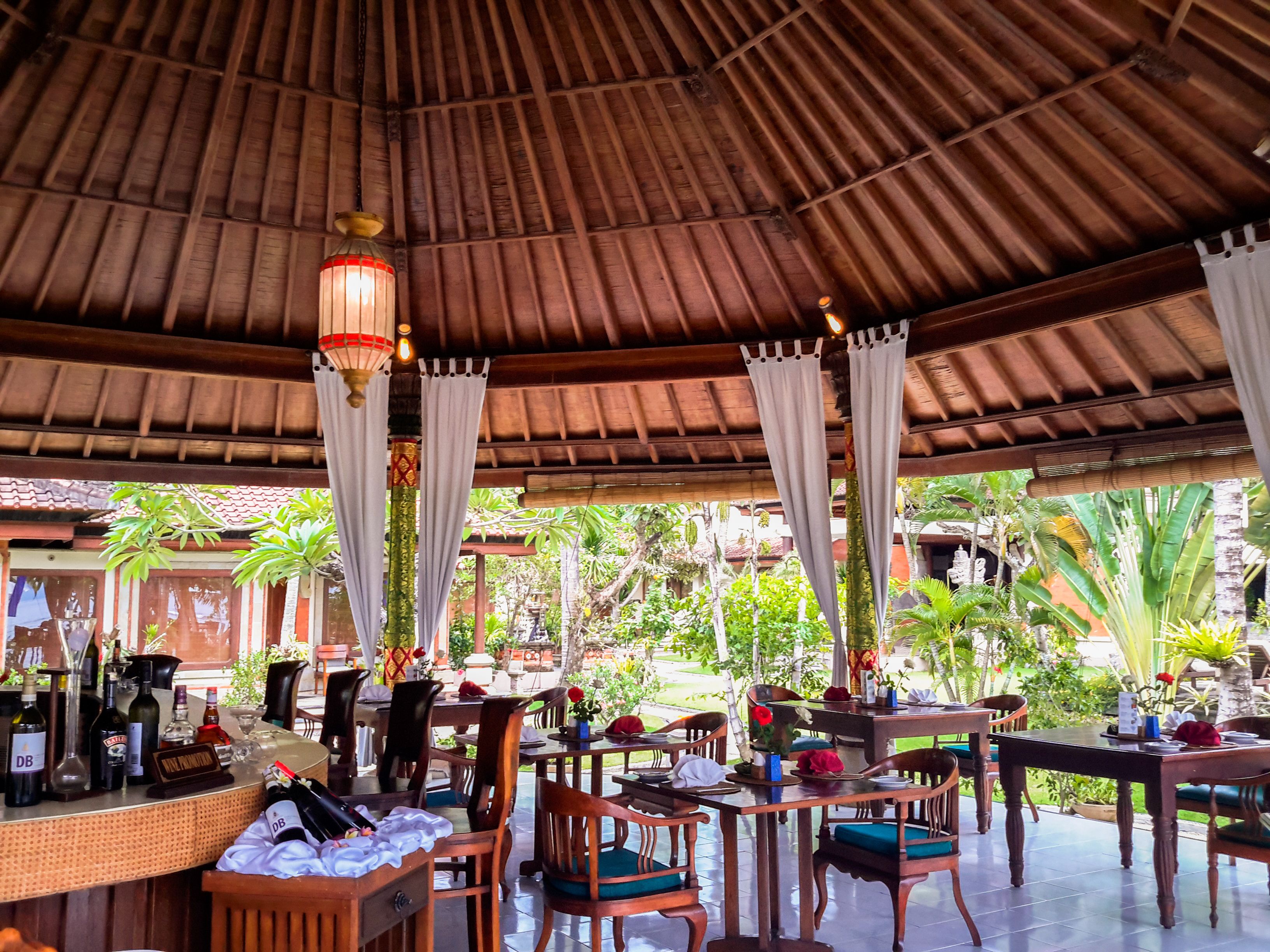An outdoor restaurant with wooden tables and chairs, a bar area with bottles, and a thatched roof.