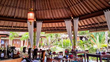 An outdoor restaurant with wooden tables and chairs, a bar area with bottles, and a thatched roof.