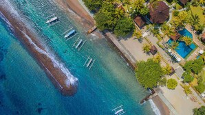 Vista aérea de un resort en la playa con aguas turquesas cristalinas, varias embarcaciones amarradas y una piscina rodeada de palmeras y protegida por estructuras de techo de paja.