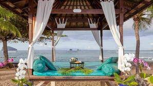 Un salón junto a la playa con un banco acolchado, una mesita con una bebida y orquídeas en macetas.