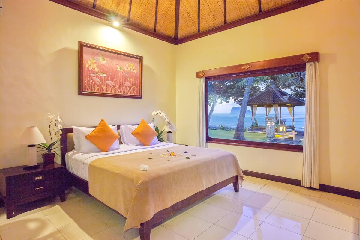 Ein helles Schlafzimmer mit einem großen Bett, einem Nachttisch, einer Topfpflanze und einem Fenster mit Blick auf einen Strand und ein Gazebo.