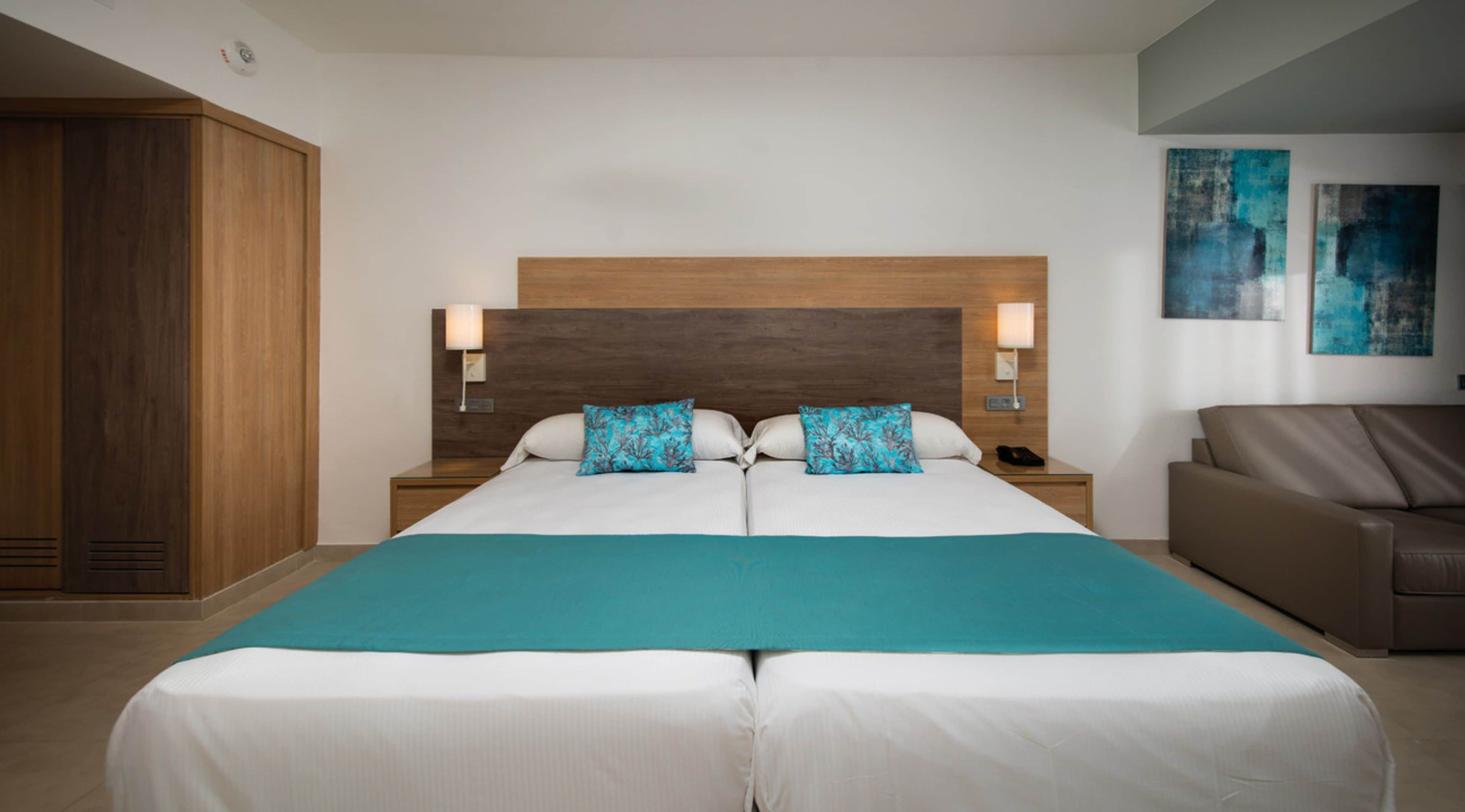Un lit double avec une couvre-lit de couleur turquoise, deux oreillers assortis, une tête de lit en bois, des lampes de chevet, un canapé en cuir marron et des œuvres d’art abstraites accrochées au mur.