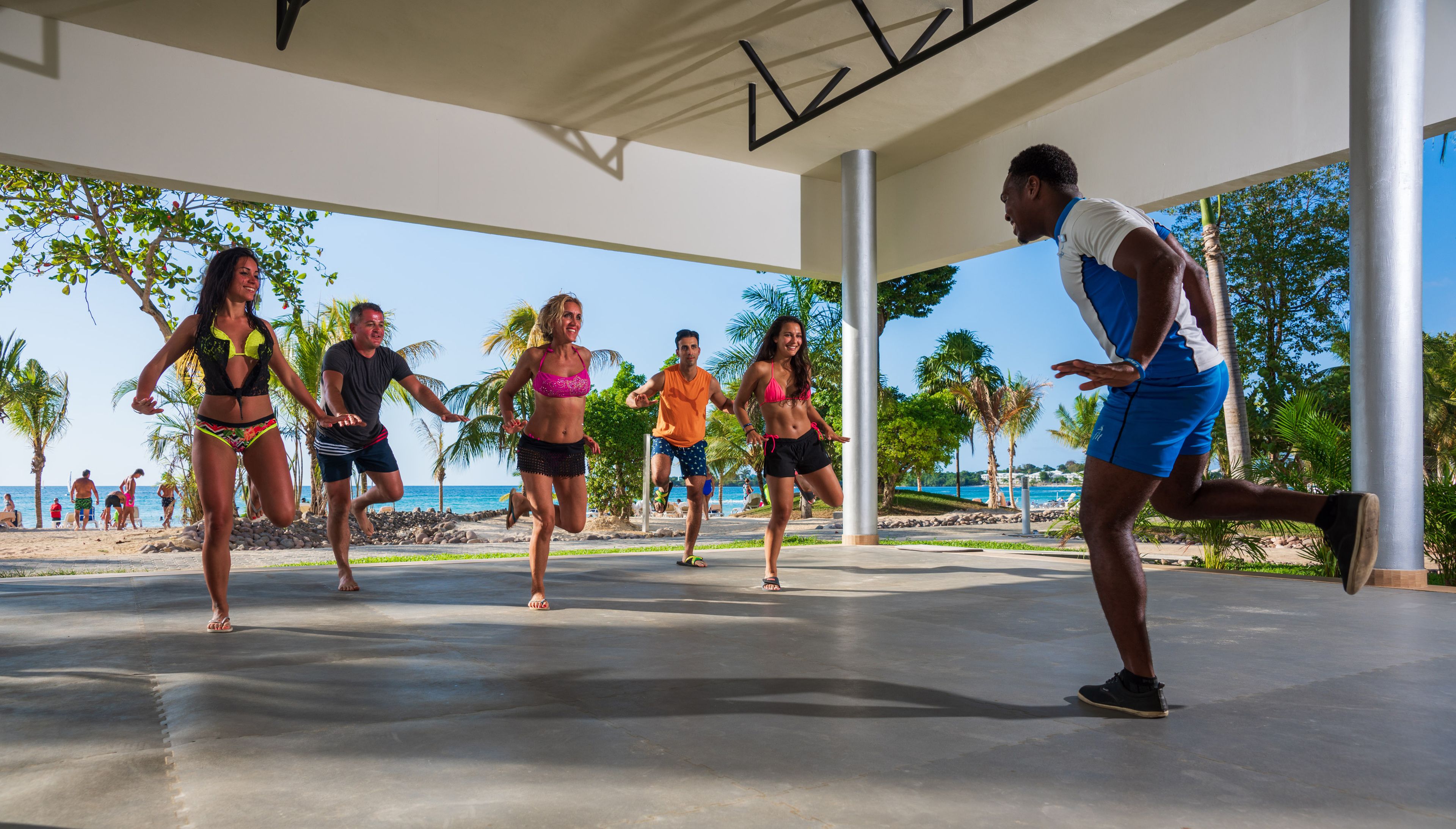Un grupo de personas haciendo ejercicio al aire libre en un espacio cubierto, con vistas a la playa y palmeras.