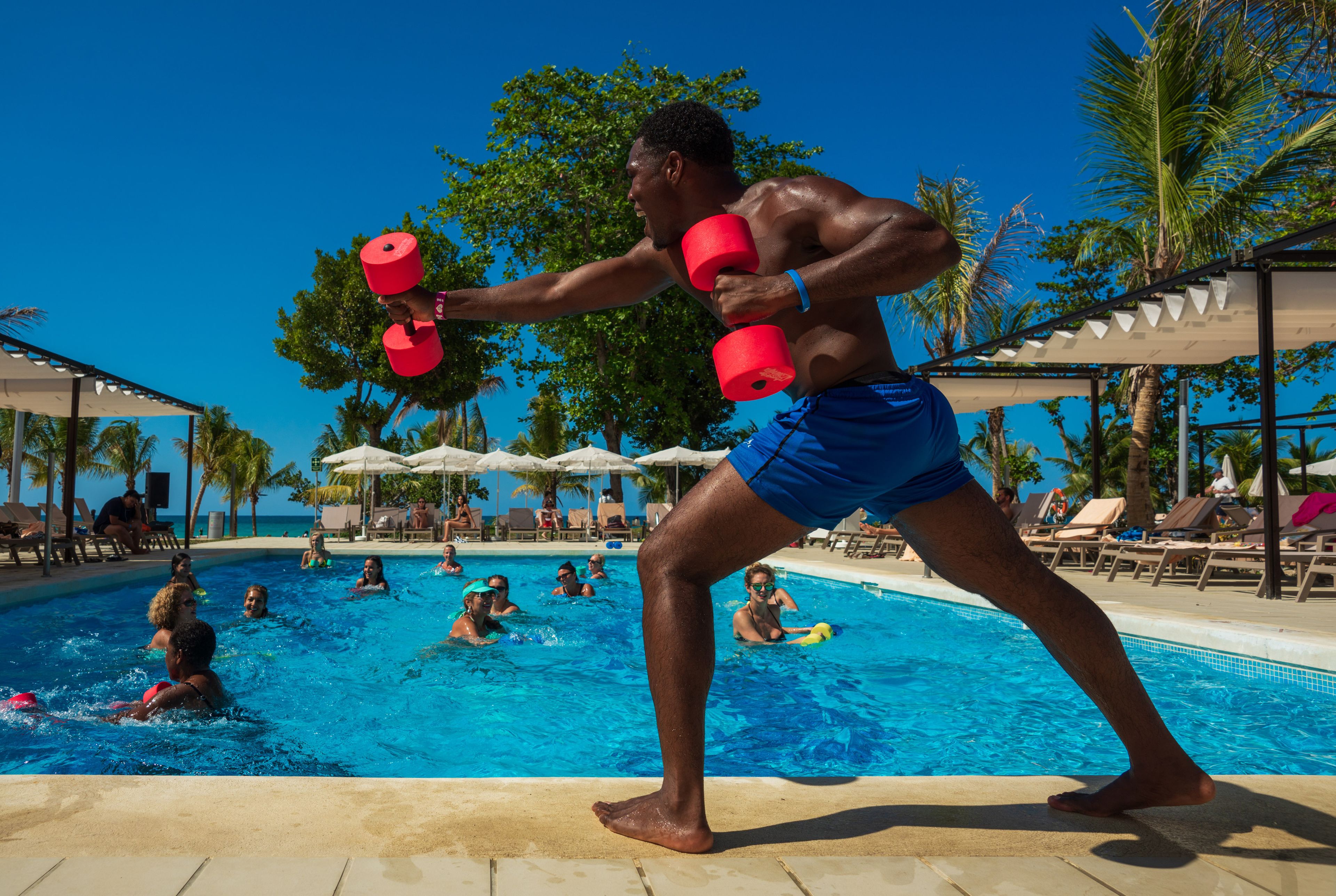 A man exercising with red dumbbells at a poolside with people swimming in the background.