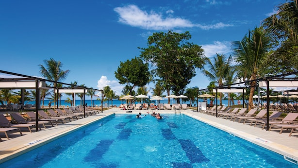A swimming pool with lounge chairs and umbrellas, surrounded by palm trees and a clear blue sky. - Hotel Riu Palace Tropical Bay - All Inclusive (Negril)