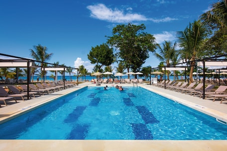 A swimming pool with lounge chairs and umbrellas, surrounded by palm trees and a clear blue sky.