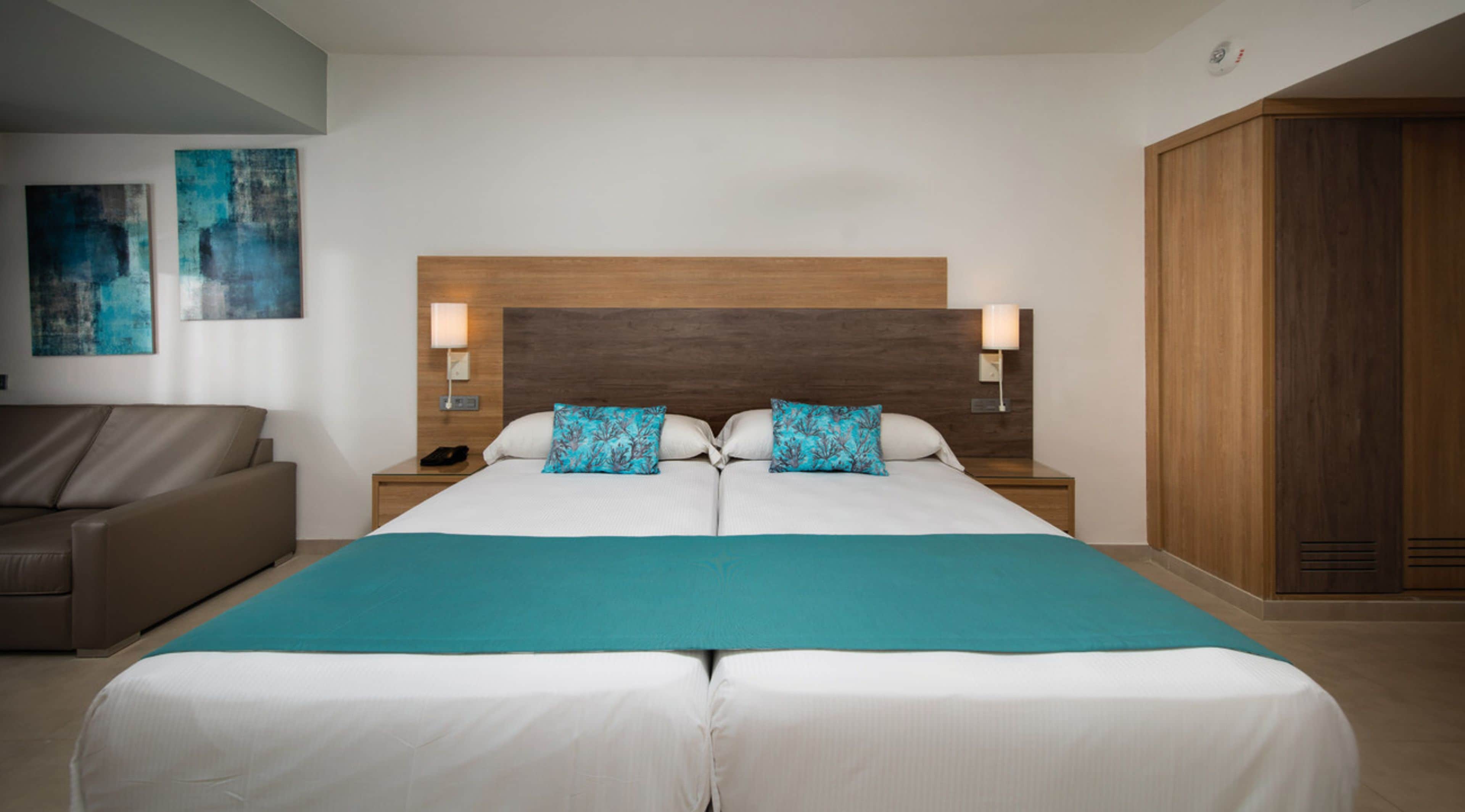 A double bed with a wooden headboard, two nightstands with lamps, a teal throw blanket, and a brown leather sofa.