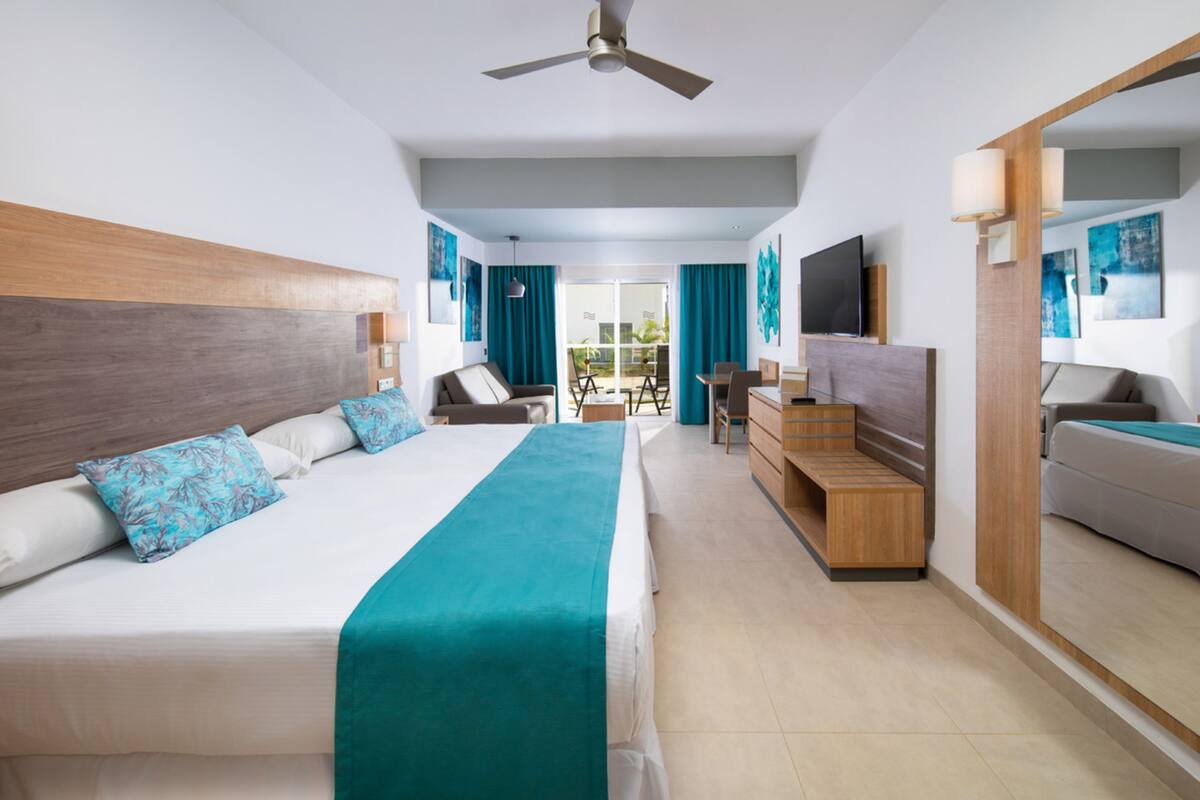 Ein Hotelzimmer mit einem großen Bett, einem Schreibtisch, einem Fernseher und einem Balkon mit blauen Vorhängen.