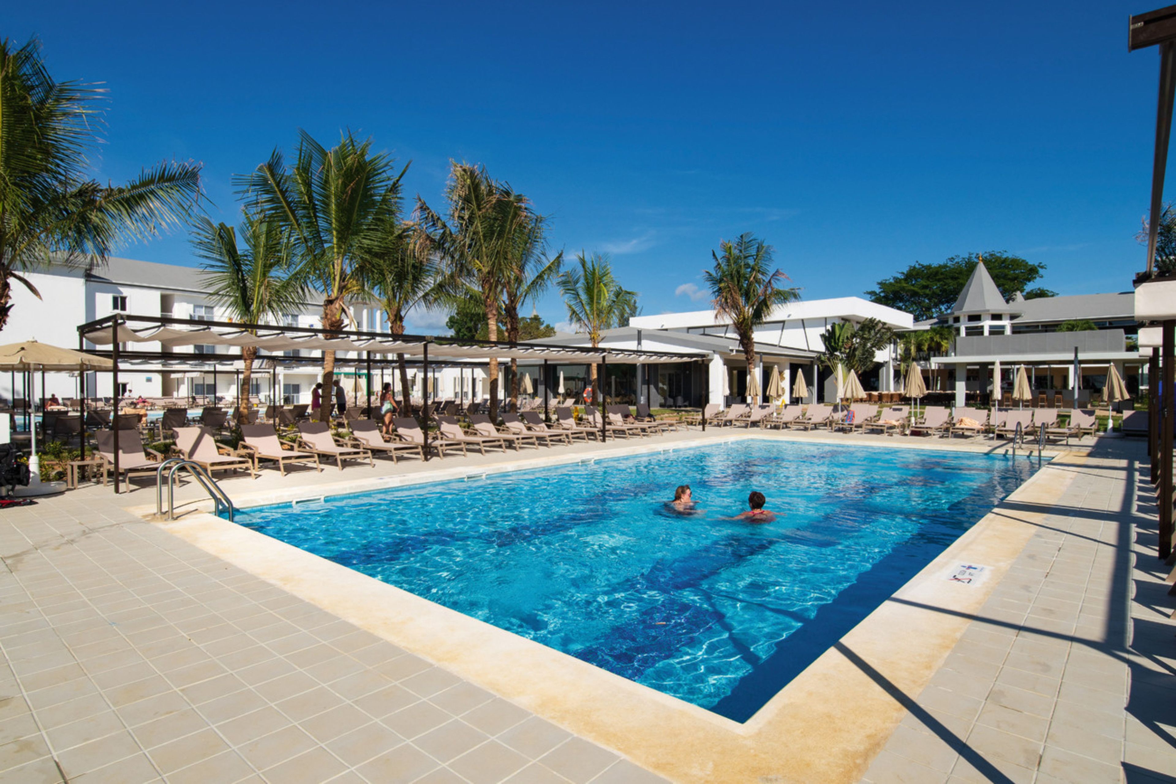 A swimming pool with lounge chairs and palm trees, surrounded by a building with a covered patio area.
