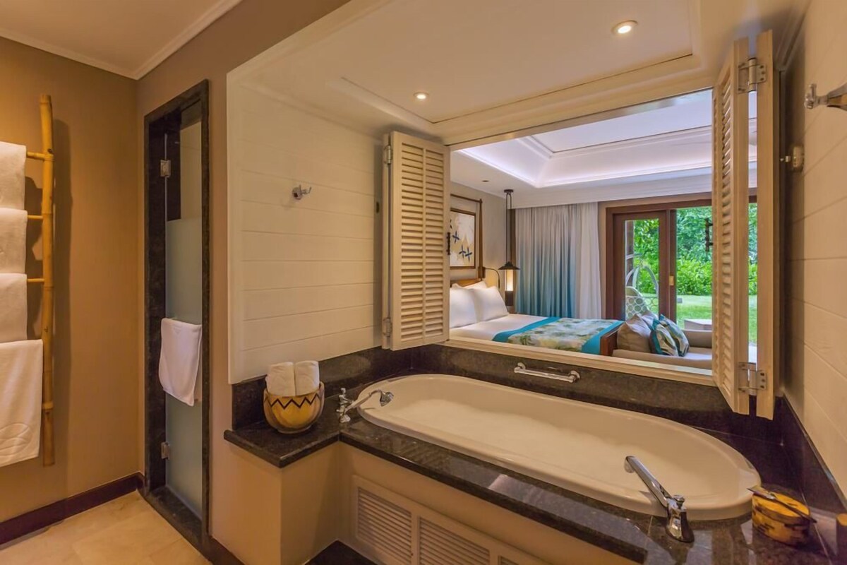 A bathroom with a large bathtub, a mirror, and a view of a bedroom with a bed and curtains.