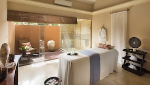 A bedroom with a large bed, a glass-enclosed shower, a sitting area with a chair and table, and a view of the outdoors.