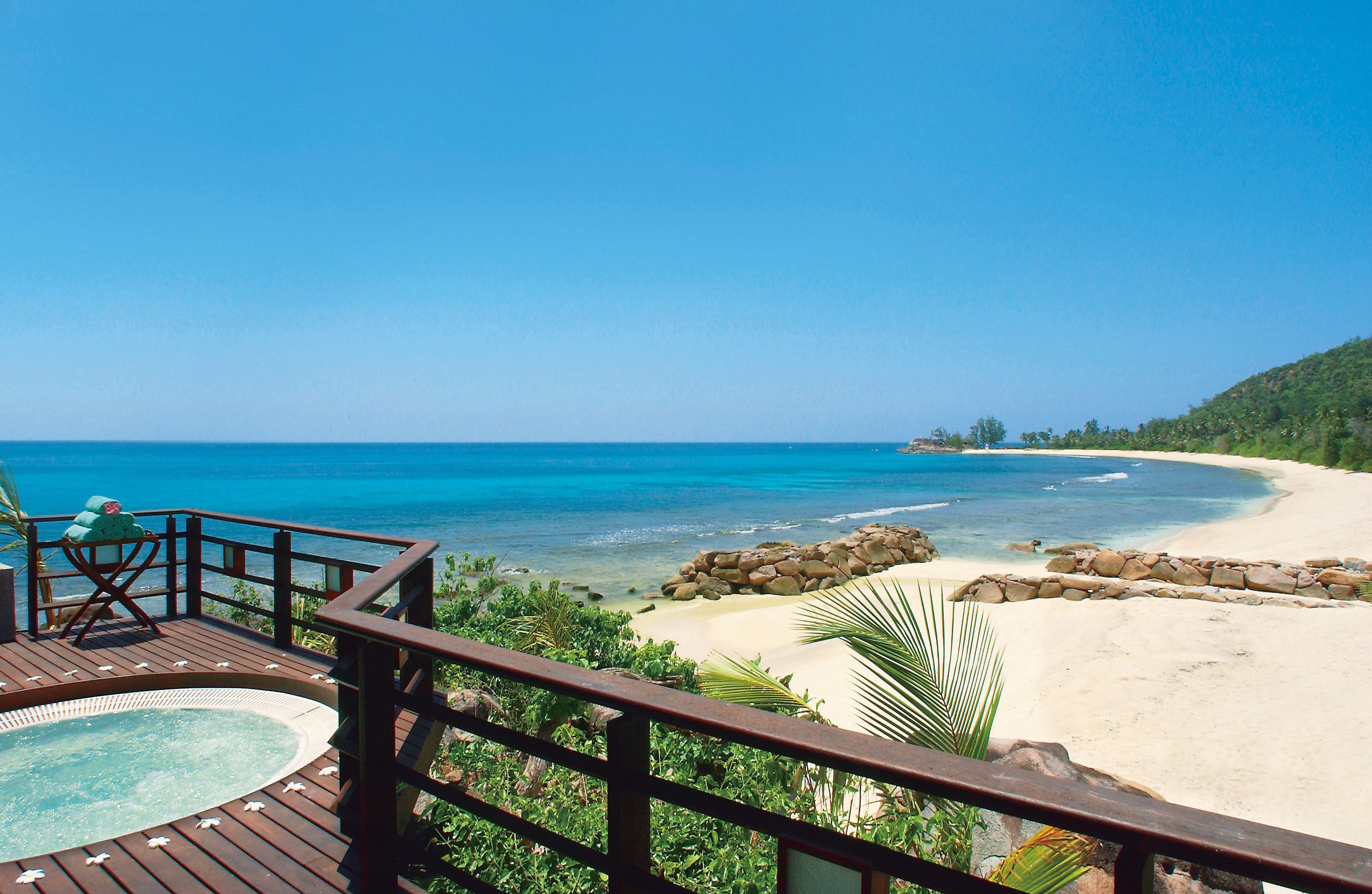 Terraza de madera con jacuzzi, con vistas a una playa de aguas azules cristalinas y arena dorada.