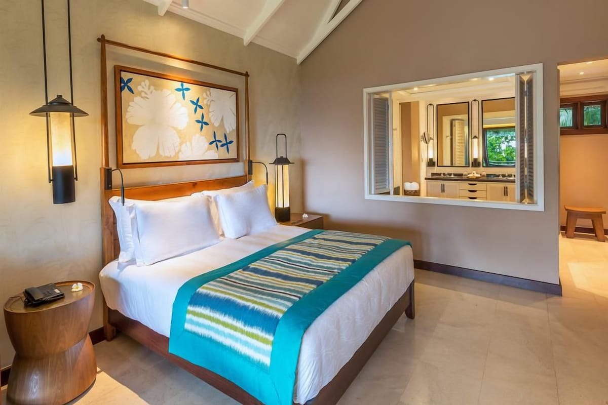 A hotel room with a large bed, a nightstand, a wooden side table, a wall-mounted mirror, and a painting above the bed.