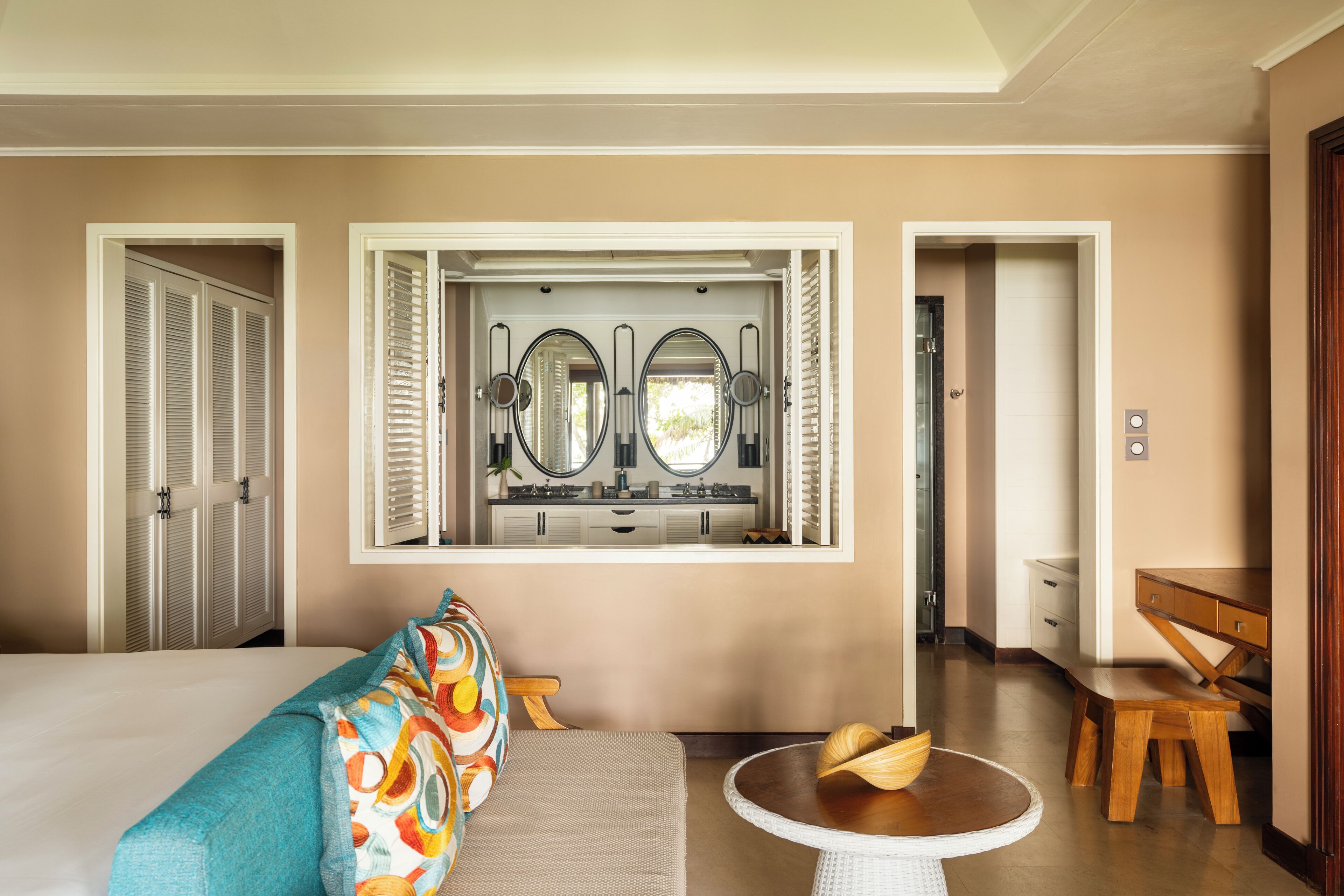 A hotel room with a bed, a sofa, a small round table, a wooden bench, and a view of a kitchen through a window.