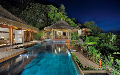 A pool area with a wooden deck, a thatched-roof structure, and lush greenery.