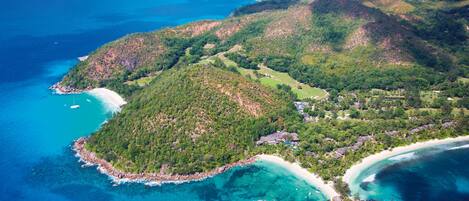 An aerial view of a coastal area with a beach, clear blue waters, and lush green hills.