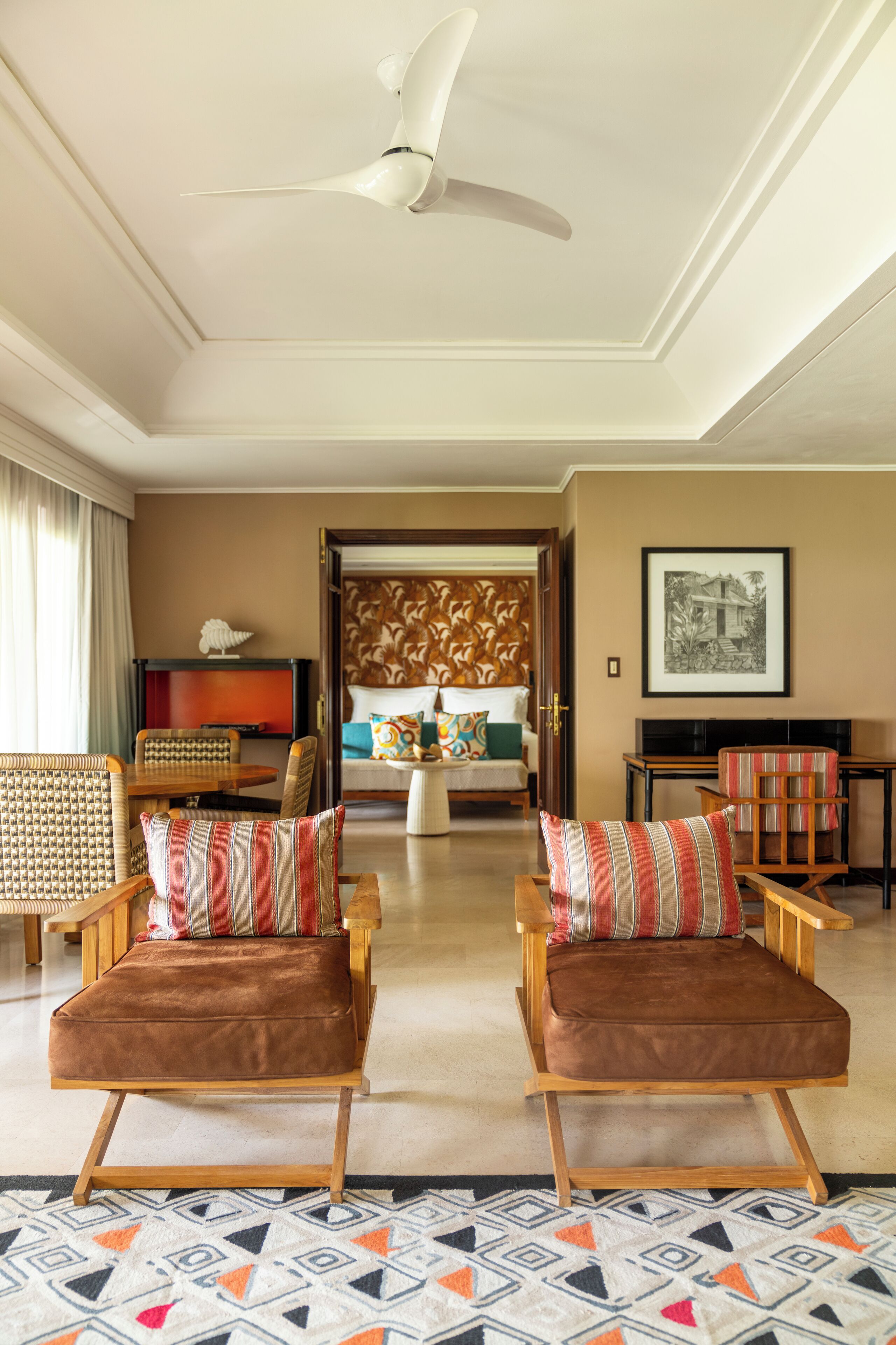 A hotel room with two wooden armchairs with brown cushions, a patterned rug, a ceiling fan, a television, and a framed picture on the wall.
