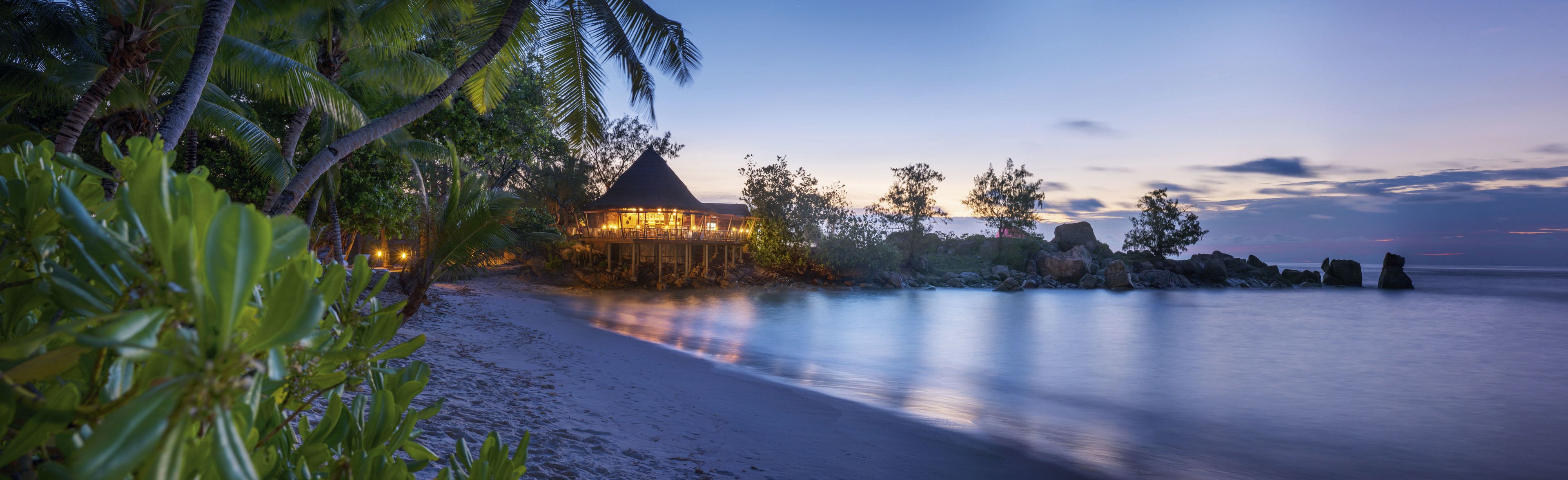 Un resort en la playa con una construcción de techo de paja, palmeras y un mar tranquilo.