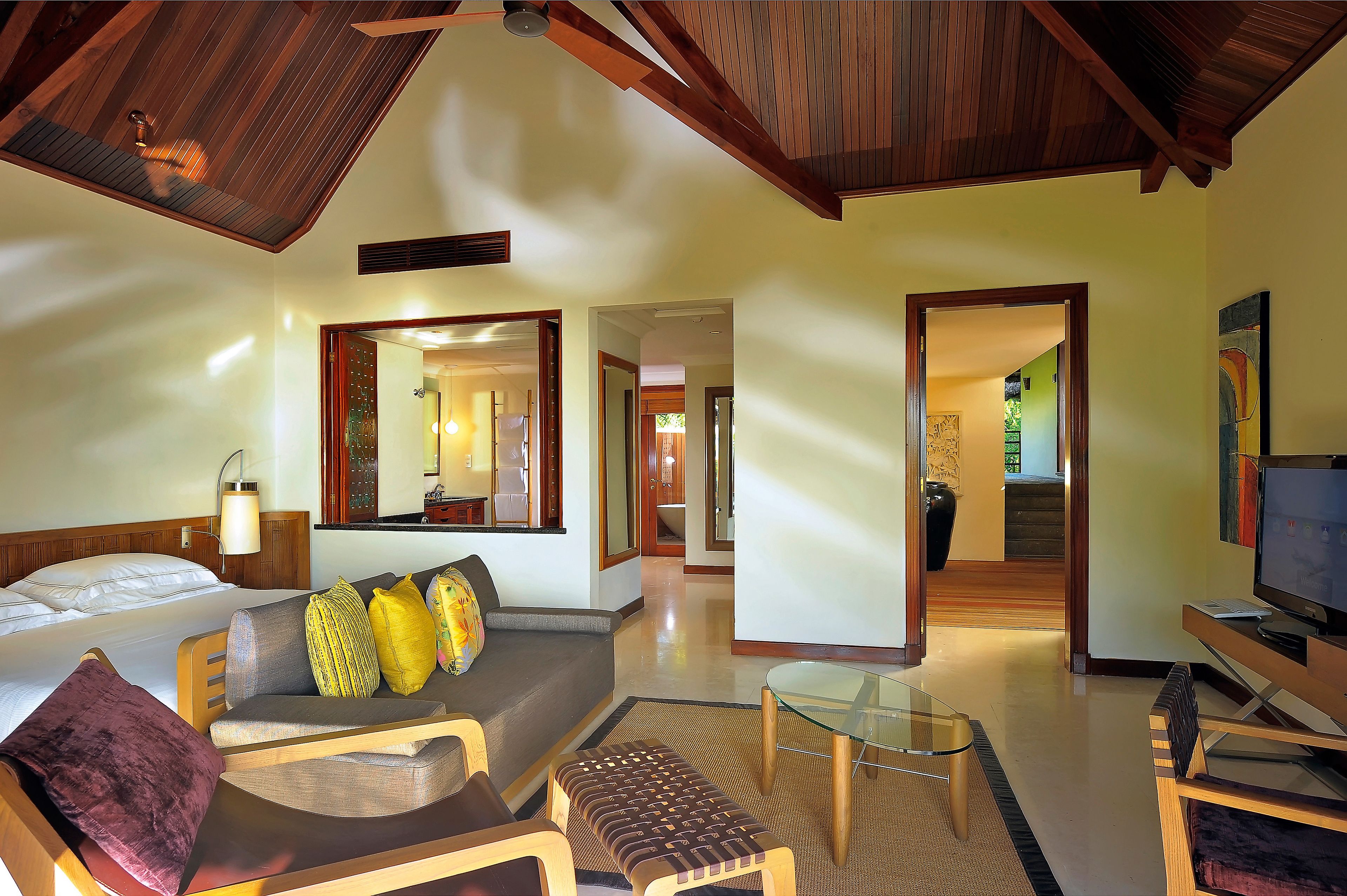 A hotel room with a bed, a sofa with cushions, a glass coffee table, a TV, and wooden beams on the ceiling.