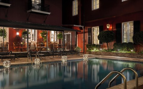 An outdoor pool area with lounge chairs and tables, surrounded by a building with large windows and a well-lit patio.