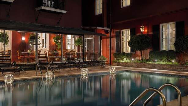 An outdoor pool area with lounge chairs and tables, surrounded by a building with large windows and a well-lit patio.