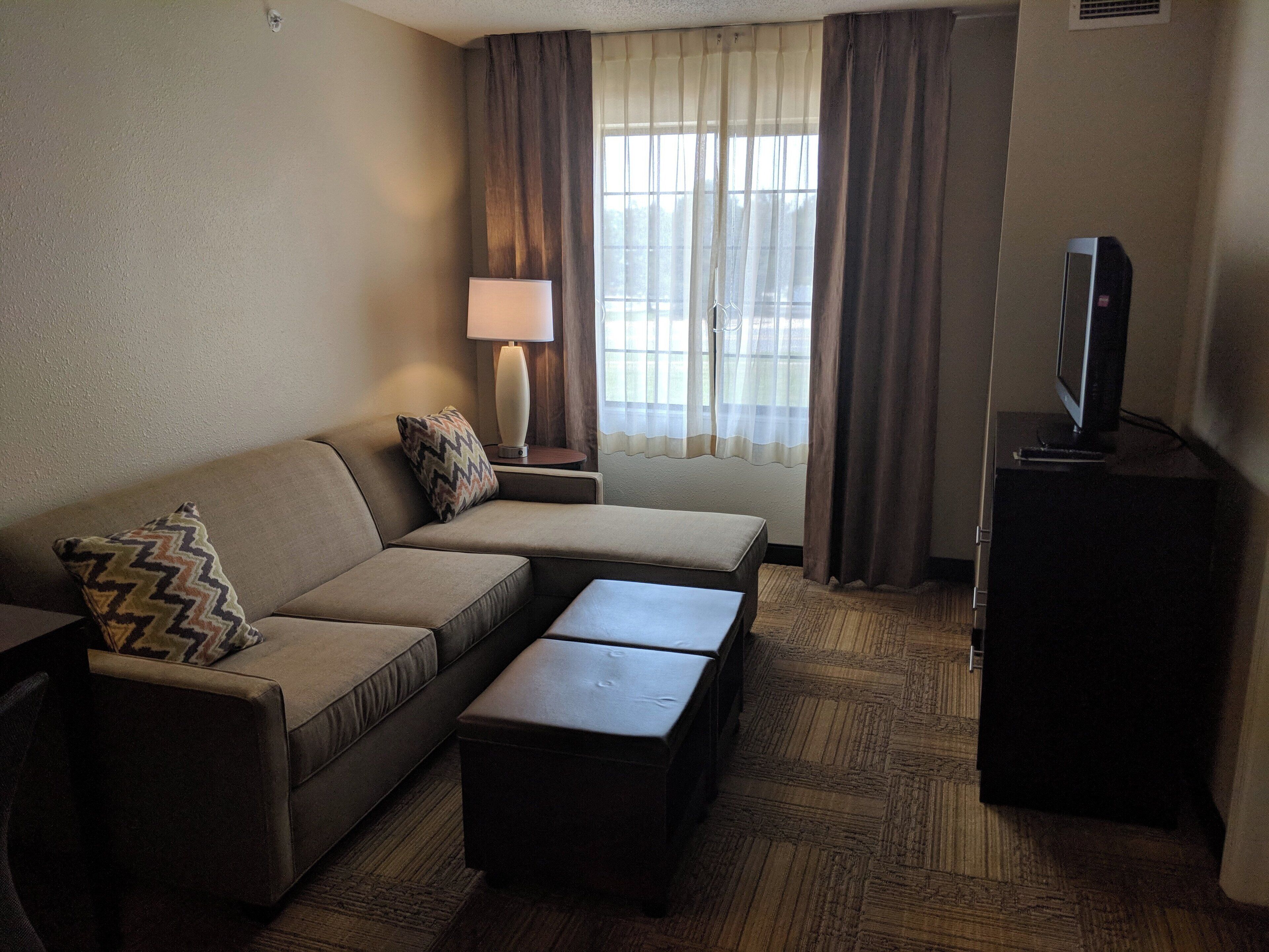 Une chambre d’hôtel avec un canapé, une petite table, une lampe, une télévision et une fenêtre avec des rideaux.